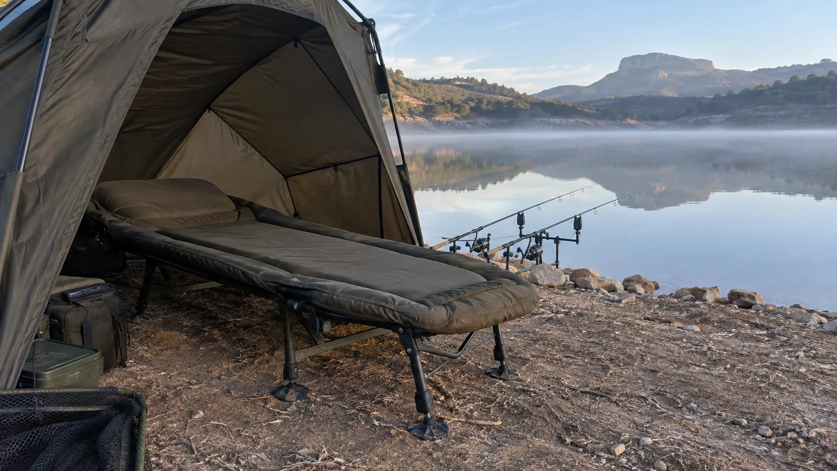 Bedchair de carpfishing montado en una orilla tranquila de embalse al amanecer.